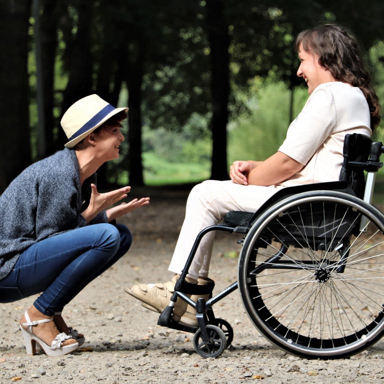 A squatting and smiling woman in front of a woman in a wheelchair, on a forest path